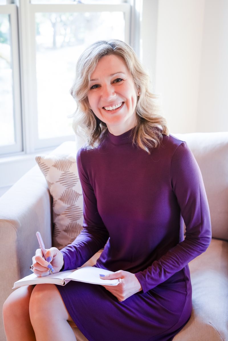 Dasha seated on a couch in a purple dress, writing in a notebook and smiling