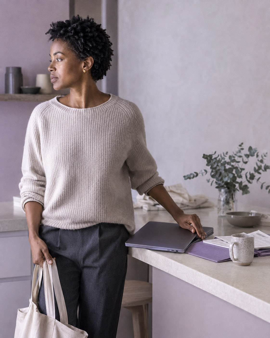 A woman closing her laptop at a kitchen counter, tote bag already in hand, on her way out